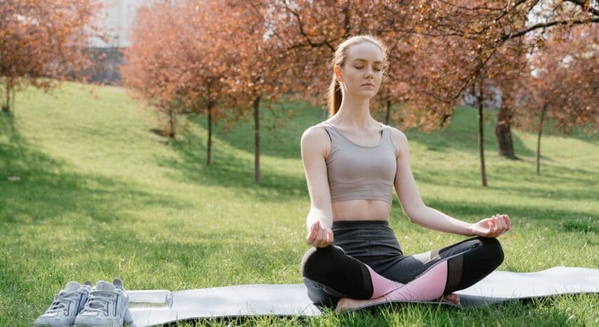 A young woman is seated on a yoga mat in a serene outdoor setting, surrounded by trees with reddish foliage. She is in a meditative pose, with her legs crossed and hands resting on her knees, eyes closed in concentration. She wears a light gray sports bra and dark leggings with pink accents. To her side, there are a pair of gray athletic shoes and a water bottle on the grass. The scene conveys a sense of tranquility and mindfulness.