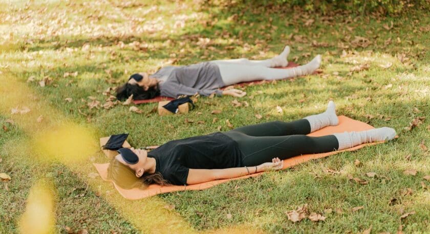 Two women lying on yoga mats outdoors on grass, wearing eye masks and comfortable clothing, appearing to be relaxing or meditating. One woman is on an orange mat wearing black leggings and a black top, while the other is on a red mat wearing white leggings and a gray top. Both have socks on and are surrounded by scattered dry leaves.