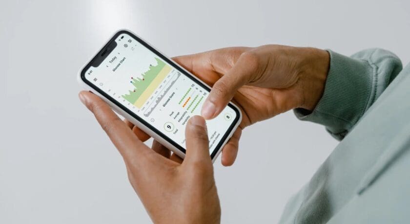 A person is holding a smartphone displaying a glucose monitoring app. The screen shows a glucose chart with various colored peaks and a score indicator. The app includes metrics such as peak, average, and total glucose levels, along with a visual representation of data trends. The individual’s hands are positioned on either side of the phone, with one finger poised to interact with the screen. The background is a light, neutral color.