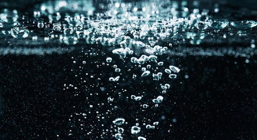 Bubbles rise through dark water, creating a dynamic scene filled with shimmering reflections. The surface is disturbed, with small droplets and bubbles visible against the contrasting dark background, enhancing the sense of movement and depth in the water.
