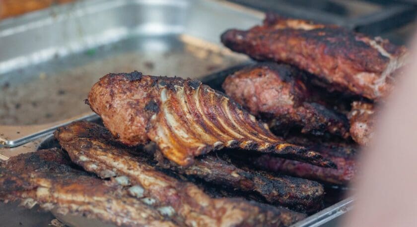 Grilled and charred pork ribs stacked on a metal tray, showing a crispy, smoky exterior with visible rib bones.