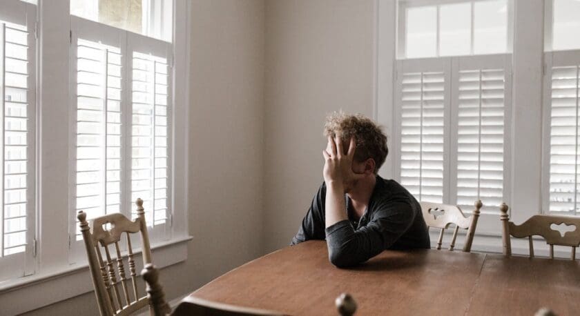 A young man sits at a wooden table, resting his head on his hand in a contemplative pose. He has curly hair and is wearing a dark shirt. The room is bright, with natural light coming through large windows adorned with white shutters. There are several empty wooden chairs around the table, and the walls are painted in a light color, creating a calm atmosphere.