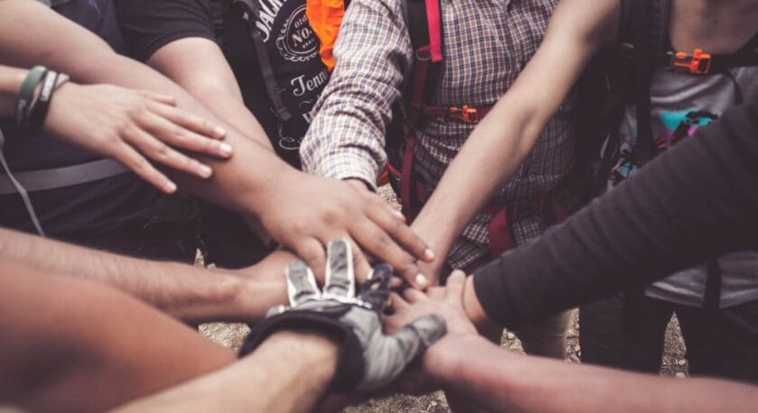 Several people with backpacks place their hands together in the center, showing teamwork or unity. One person is wearing a glove, and the group appears to be outdoors.