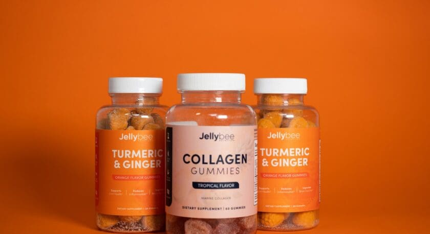 Three clear plastic bottles of dietary gummies are displayed against a bright orange background. Two bottles on the sides contain "Turmeric & Ginger" gummies in orange flavor, while the center bottle is labeled "Collagen Gummies" in tropical flavor. Each bottle features a colorful label with the brand name "Jellybee" and highlights the benefits of the supplements. The gummies inside are visible, showcasing their vibrant colors.