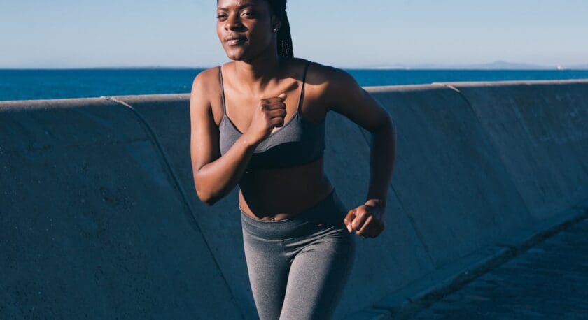 A woman jogging outdoors near a concrete barrier with the ocean in the background. She is wearing a grey sports bra and grey leggings, with her hair pulled back. The sky is clear and the lighting suggests it is either early morning or late afternoon.