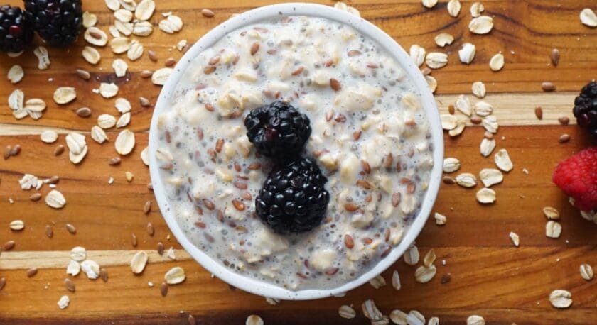 A white bowl filled with overnight oats mixed with chia seeds and flaxseeds, topped with two blackberries, placed on a wooden surface scattered with oats, flaxseeds, blackberries, and a raspberry.