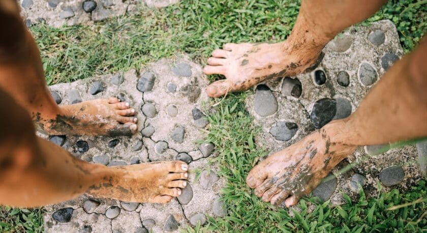 Two pairs of feet are shown on a stone pathway surrounded by grass. One pair has muddy feet with visible dirt and some grass stains, while the other pair has cleaner feet with painted toenails. The stones in the pathway are irregularly shaped, and the grass is lush and green, contrasting with the mud on the feet.