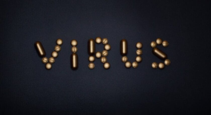 The image features the word "VIRUS" creatively arranged using various golden-colored capsules and spherical shapes. The background is a solid black, which enhances the visibility of the golden elements. The letters are formed by a combination of elongated capsules and round shapes, creating a visually striking contrast.