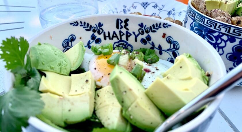 A bowl filled with sliced avocado, a poached egg, and chopped green onions sits on a table. The bowl features a decorative blue and white pattern with the phrase "BE HAPPY" written inside. Fresh cilantro is visible alongside the avocado. In the background, a glass of water and another bowl containing what appears to be roasted potatoes can be seen, along with a drink in a glass. The setting suggests a casual dining experience.