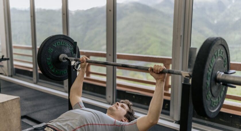 A person is performing a bench press in a gym setting, lying on a bench with a barbell held above their chest. The barbell is loaded with black weight plates. The background features large windows that offer a view of green mountains, creating a scenic atmosphere. The individual is wearing a fitted gray shirt and appears focused on the lift. The gym floor is covered with mats, and there is a wooden box nearby.