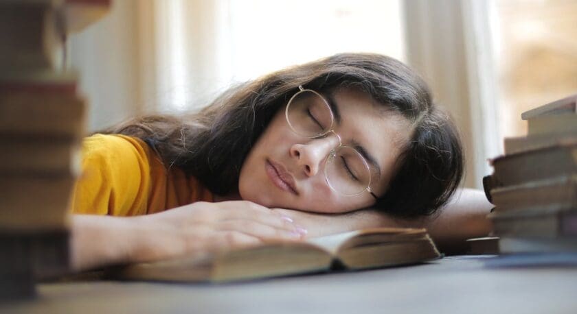 A young person with long, dark hair is resting their head on an open book, eyes closed, appearing peaceful and relaxed. They are wearing round glasses and a yellow shirt. Surrounding them are stacks of books, creating a cozy, studious atmosphere. Soft natural light filters in through nearby windows, enhancing the serene setting.