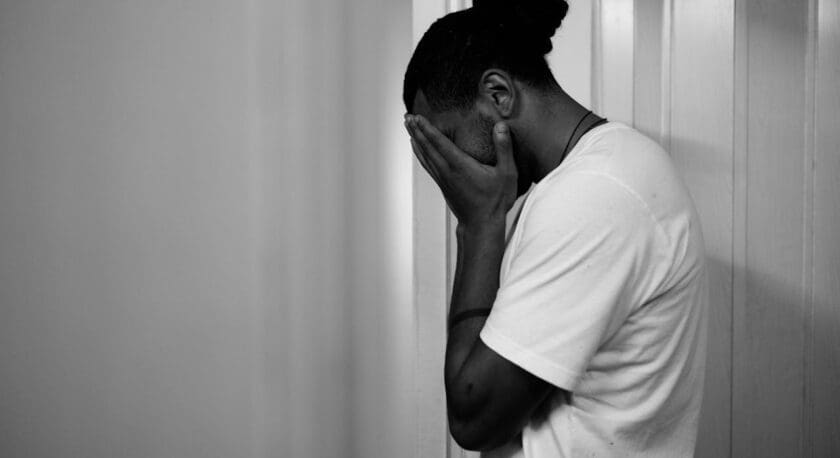 A person stands against a wall, facing slightly away. They are wearing a plain white t-shirt and have their hands covering their face, suggesting a moment of distress or contemplation. The image is in black and white, emphasizing the emotional tone. The background features a simple, light-colored wall with vertical paneling.