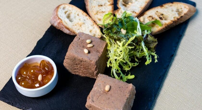 Two rectangular pieces of pâté garnished with pine nuts, served on a black slate plate with mixed greens, four slices of toasted baguette, and a small white bowl of amber-colored jelly.