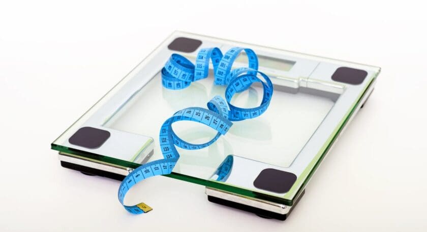 A glass bathroom scale is positioned on a light background. A blue measuring tape is coiled on top of the scale, displaying various weight measurements. The scale features a sleek design with black rubber feet and a digital display area.