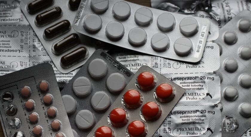 Several blister packs of various pills and capsules are shown, including round gray tablets, red round pills, and dark brown capsules. Some packaging displays text indicating "Loseprazol 20 mg" and "omeprazolum," suggesting these are medications. The packs are overlapping on a flat surface.