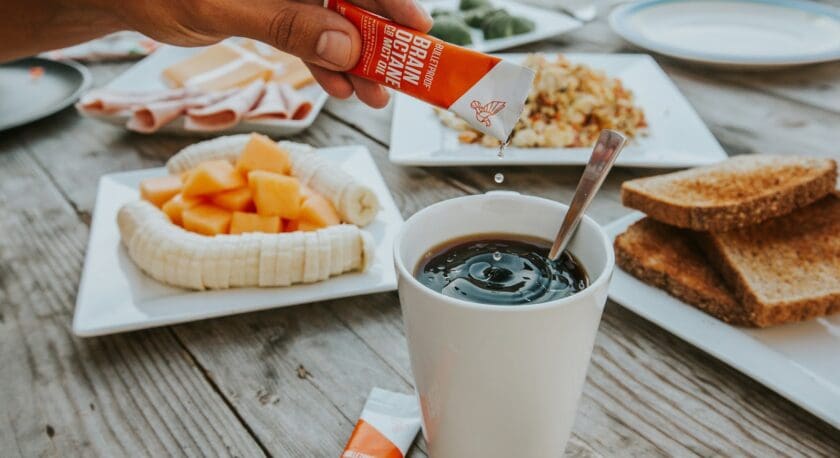 A hand is squeezing a packet of Bulletproof Brain Octane C8 MCT Oil into a white cup filled with a dark liquid, likely coffee. The cup has a spoon inside. Surrounding the cup on a wooden table are plates with sliced bananas and cantaloupe, toast, scrambled eggs, and slices of deli meat.