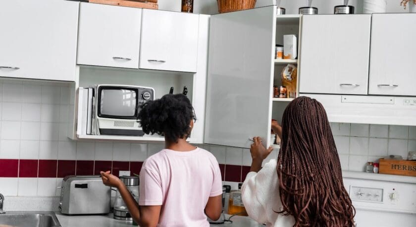 Two women are in a kitchen, one with short curly hair wearing a light pink shirt, and the other with long braided hair wearing a white top. They are standing near white cabinets, with the woman on the right opening a cabinet door. The kitchen has a countertop with a toaster, coffee maker, and a vintage-style microwave. The backsplash features white tiles with a horizontal strip of dark red tiles. There are decorative items on top of the cabinets, including a wicker basket and a brown vase with dried flowers.