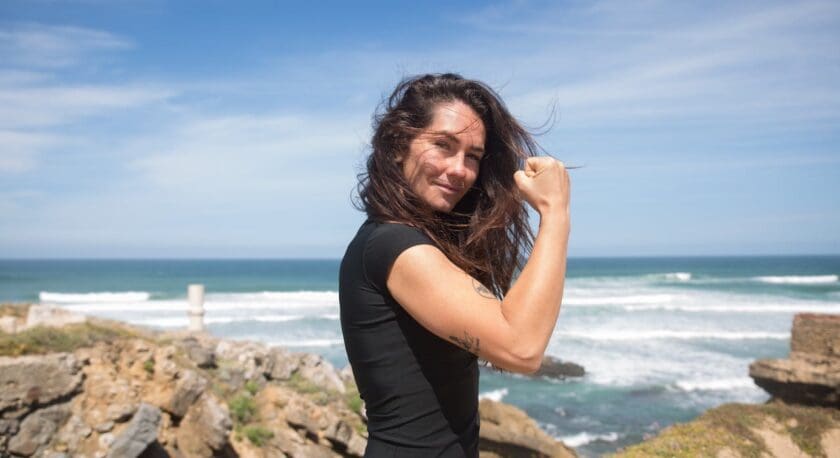 A woman with long brown hair wearing a black short-sleeve top stands outdoors near rocky cliffs by the ocean. She is flexing her right arm and smiling confidently, with waves and a blue sky in the background.
