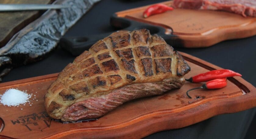 A perfectly grilled piece of meat is displayed on a wooden cutting board, showcasing a beautifully charred surface with a crisscross pattern. The meat is juicy, with a hint of pink visible at the edges. Next to it, there are two small red chili peppers and a sprinkle of coarse salt, adding a touch of color and flavor contrast. In the background, additional cuts of meat are visible on separate wooden boards. The setting has a rustic feel, with dark surfaces complementing the rich colors of the food.