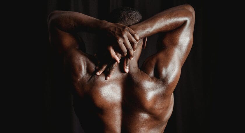 Muscular bare back of a person with dark skin, arms raised and hands clasped behind the head, set against a black background.
