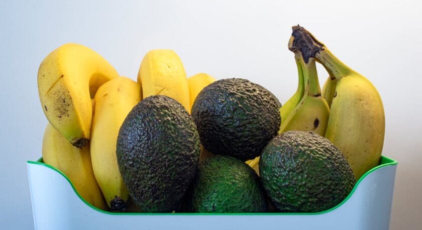 A collection of ripe bananas and avocados is arranged in a white container with a green edge. The bananas are bright yellow, some with slight blemishes, while the avocados are dark green with a textured skin. The background is plain and light, emphasizing the vibrant colors of the fruits.