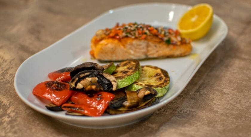 A white plate features a portion of grilled salmon topped with a colorful salsa and a lemon wedge on the side. Accompanying the salmon are grilled vegetables, including red bell peppers, zucchini, and mushrooms, arranged attractively on the plate. The dish is presented on a textured surface, enhancing its visual appeal.