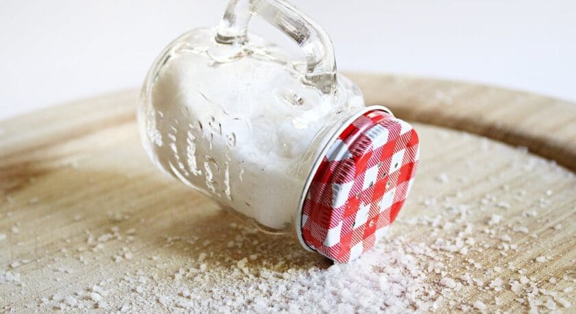 A glass jar with a red and white checkered lid is tipped over on a wooden surface, spilling white salt around it. The jar has a handle and is partially filled with salt, creating a scattered effect on the surface. The background is a soft, neutral color, emphasizing the jar and the salt.