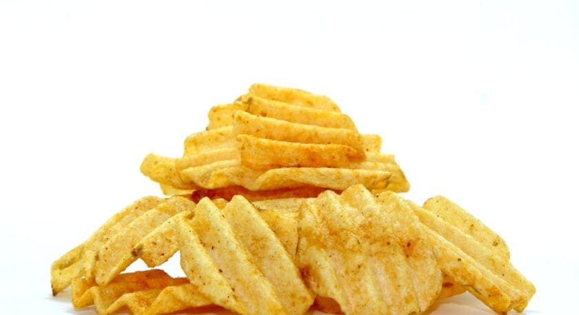 A pile of wavy potato chips is displayed against a white background. The chips are golden brown with a slightly crispy texture, showcasing ridges and some seasoning. The arrangement is casual, with some chips stacked on top of others, creating a visually appealing snack presentation.