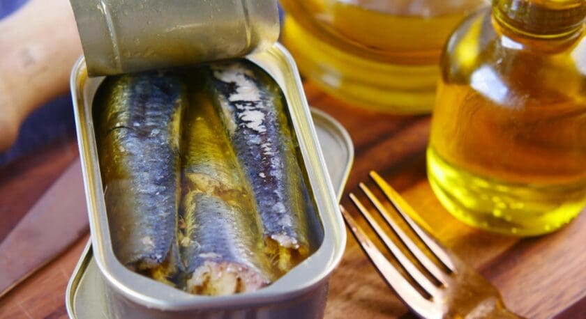 A tin can is partially opened, revealing several sardines packed in oil. The sardines are glistening and arranged neatly inside the can. Nearby, there is a fork and a bottle of olive oil, along with a wooden cutting board. The background features a blurred hand holding a knife.