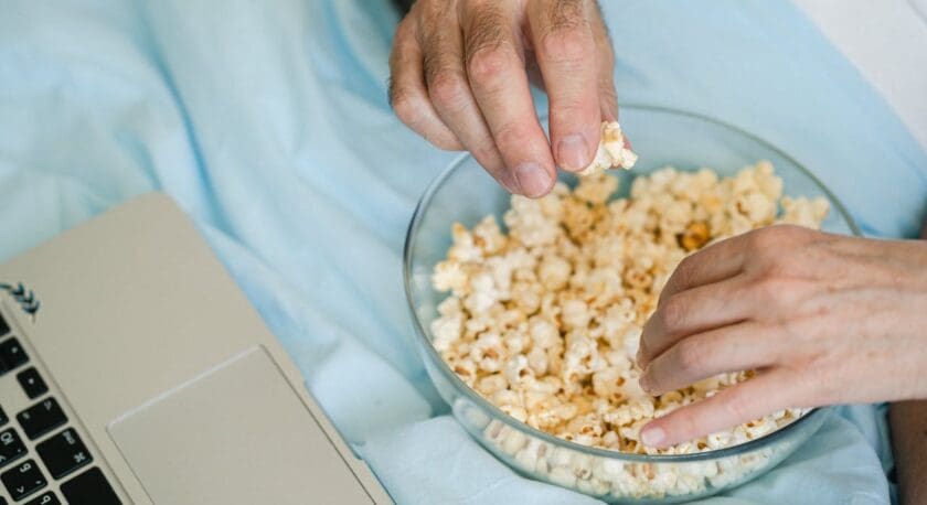 Two hands reaching into a clear glass bowl filled with popcorn, placed on a light blue fabric surface next to a silver laptop.