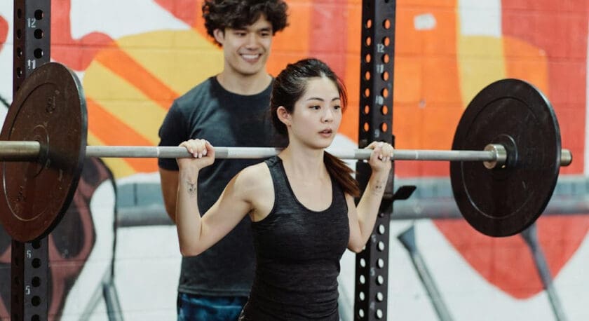 A woman is performing a weightlifting exercise, holding a barbell above her shoulders. She is wearing a black tank top and patterned leggings. Behind her, a man is smiling and providing encouragement. The background features a colorful mural on the wall, adding vibrancy to the gym setting. The gym equipment, including a squat rack, is visible on either side.