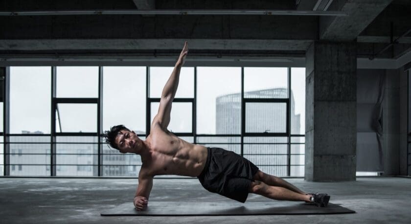 A shirtless man wearing black shorts and black shoes is performing a side plank exercise on a black mat in an industrial-style room with large windows and concrete walls. His left forearm is on the mat, supporting his body, while his right arm is extended upward.