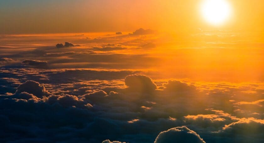 A vibrant sunset is depicted above a sea of clouds, with the sun positioned low on the horizon. The sky transitions from deep blue to warm hues of orange and yellow, illuminating the clouds below. The clouds appear fluffy and varied in shape, creating a soft, textured landscape against the colorful backdrop.