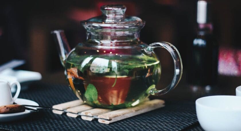 A glass teapot filled with green tea and fresh mint leaves sits on a wooden trivet. Nearby, a small white cup and a plate with a cookie are visible on a textured black placemat. The background is softly blurred, creating a cozy atmosphere.
