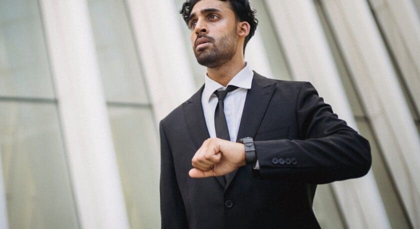A man in a formal black suit stands outdoors, looking slightly to the side with a serious expression. He is checking the time on his wristwatch, which is visible on his left hand. The background features modern architecture with tall, light-colored panels. The man's shirt is white, and he wears a black tie. His hair is dark and curly, adding to his professional appearance.