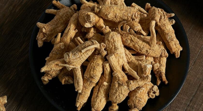 A black plate filled with several dried ginseng roots, placed on a wooden surface. The ginseng roots are light brown, wrinkled, and have a distinct, elongated shape with small offshoots.