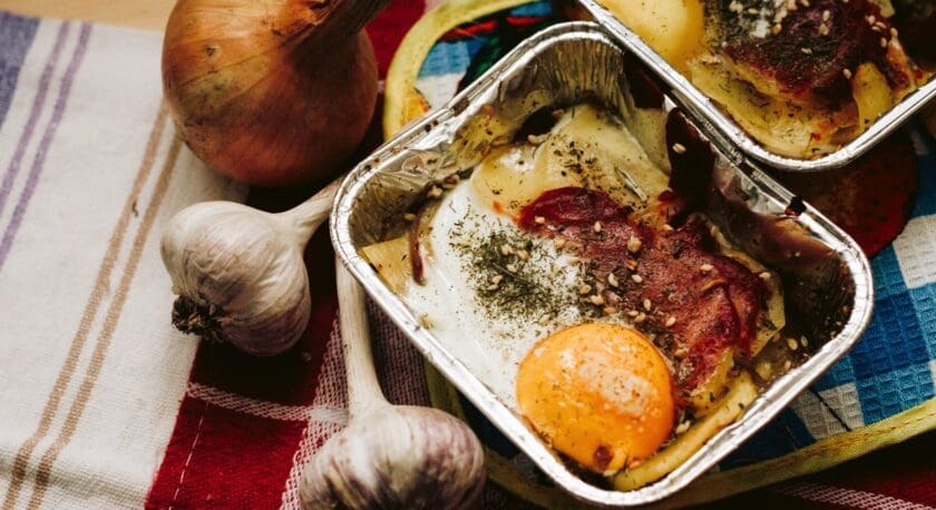 Two foil containers with baked eggs topped with herbs and slices of cured meat, placed on a colorful cloth. Nearby are a whole onion and two garlic bulbs.