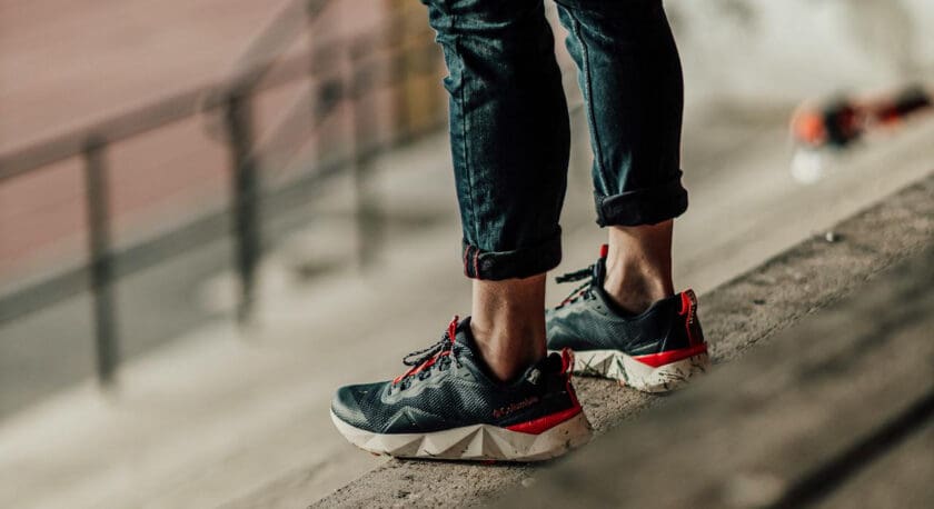 A person wearing black and red athletic shoes with white soles, standing on a concrete ledge. The person is dressed in rolled-up dark pants, and the background is blurred with a railing and some indistinct objects.