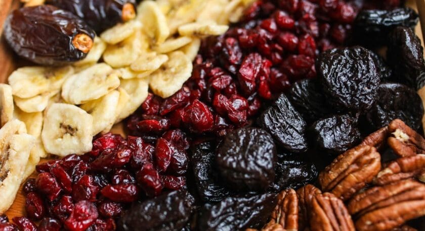 A close-up of an assortment of dried fruits and nuts, including dried dates, banana chips, dried cranberries, dried prunes, and pecans arranged closely together.