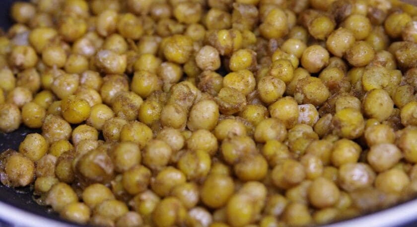 Roasted chickpeas coated with spices in a pan, showing a golden-brown color and a slightly crispy texture.