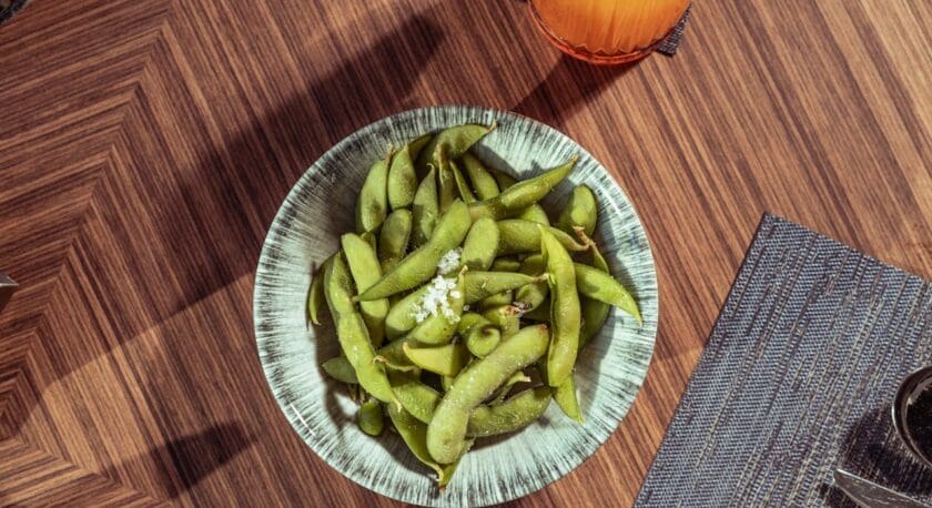 A bowl of green edamame pods sprinkled with coarse salt on a wooden table. Nearby are a glass of orange-colored drink with ice and a dark-colored cup, along with a blue woven placemat.