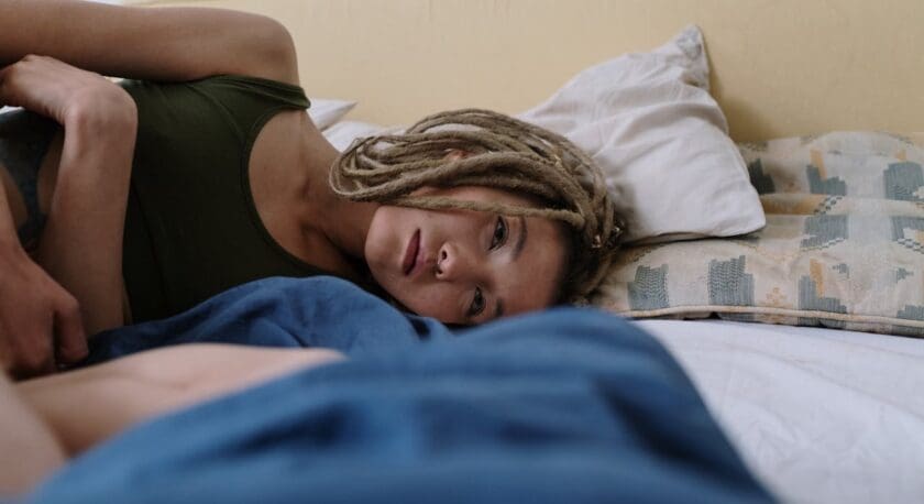 A person with light brown dreadlocks lying on a bed, resting their head on a pillow. They are wearing a dark green sleeveless top and looking ahead with a calm expression. The bed has a white sheet and a patterned pillow behind the person.