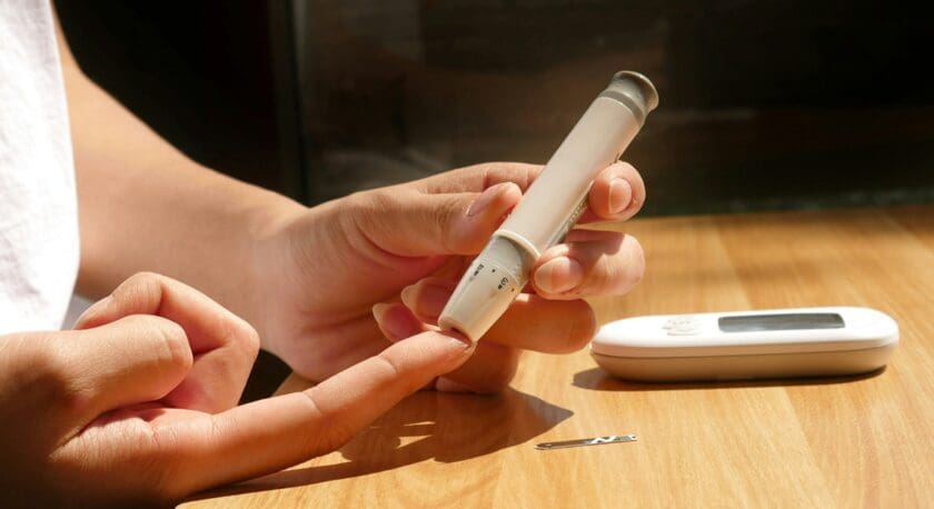 Hands using a lancet device to prick a finger for a blood glucose test, with a glucose meter and a lancet needle on a wooden surface.