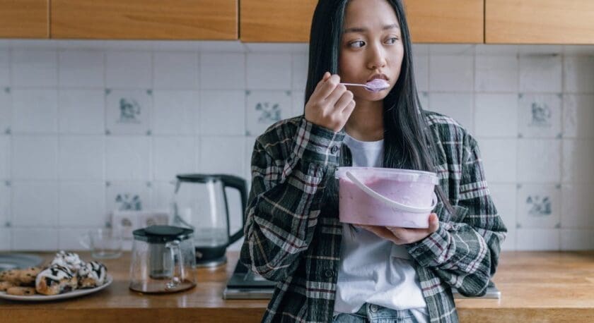 A young woman with long dark hair wearing a plaid shirt over a white t-shirt and jeans is standing in a kitchen. She is holding a large plastic container filled with pink yogurt or a similar creamy food and eating from it with a spoon. The kitchen has wooden countertops, white tiled walls with some decorative tiles, and various items including a glass kettle, a coffee pot, and a plate of pastries.
