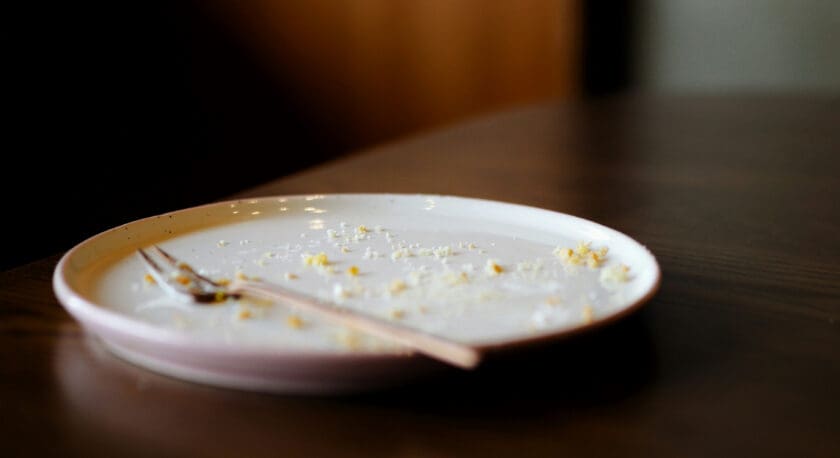 A white plate with crumbs scattered on it and a fork resting on the edge, placed on a dark wooden table.