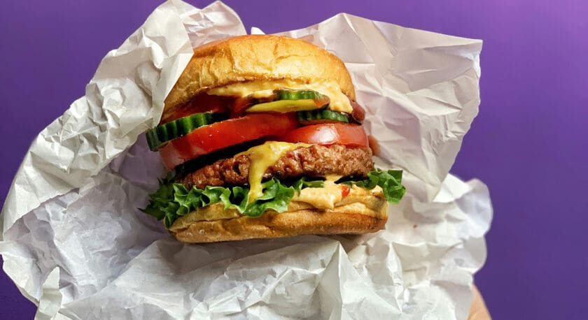 A hand holding a wrapped burger with a toasted bun, lettuce, a beef patty, melted cheese, tomato slices, cucumber slices, and sauce, against a purple background.