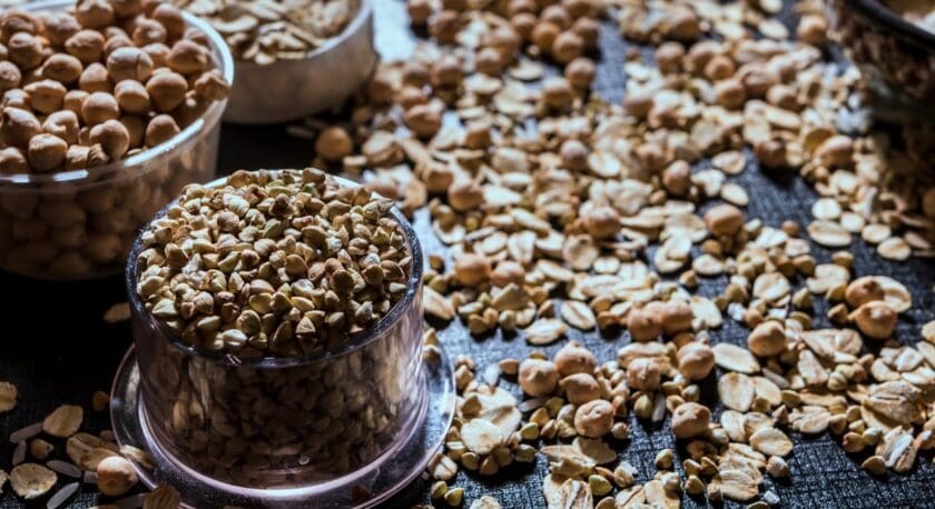 A close-up of various grains and seeds scattered on a dark textured surface, with some contained in clear plastic containers. The grains include oats, chickpeas, and buckwheat. The lighting highlights the textures and natural colors of the grains.