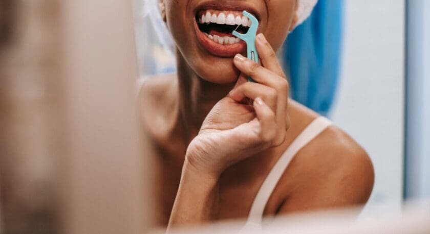 Close-up of a person using a blue dental floss pick to clean their teeth, with a white towel wrapped around their head and wearing a white tank top.