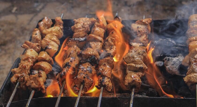 Skewers of marinated meat grilling over an open flame with visible flames and smoke, showing charred and cooked pieces on a barbecue grill.