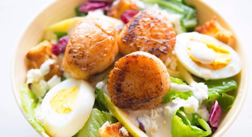 A salad bowl featuring three seared scallops on top, halved hard-boiled eggs, mixed greens, croutons, dried cranberries, and a creamy dressing.